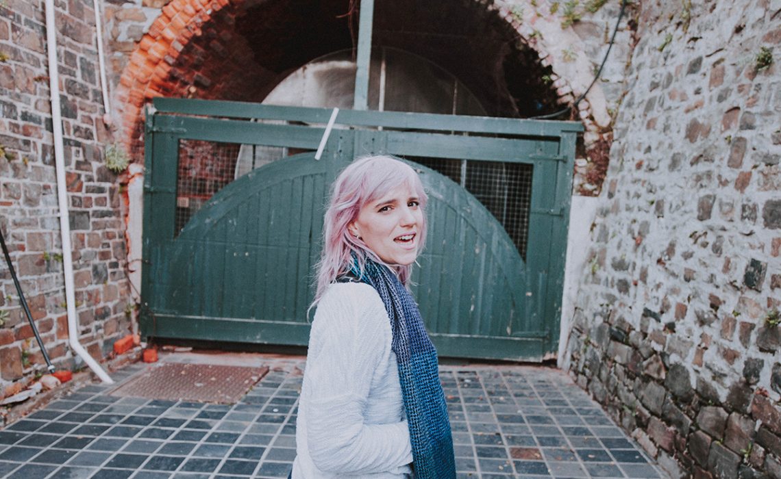 Girl with purple hair, standing in front of large door.